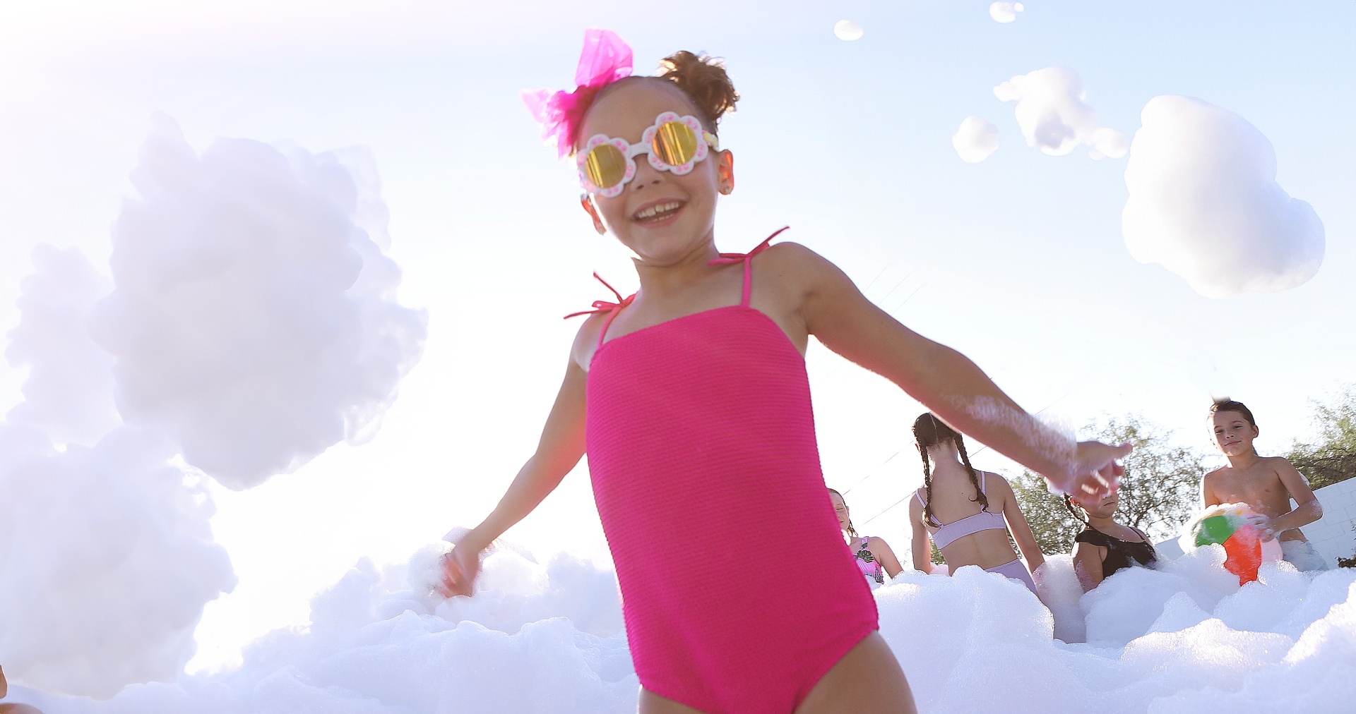 Little girl in pink swimsuit and sunglasses dancing in foam